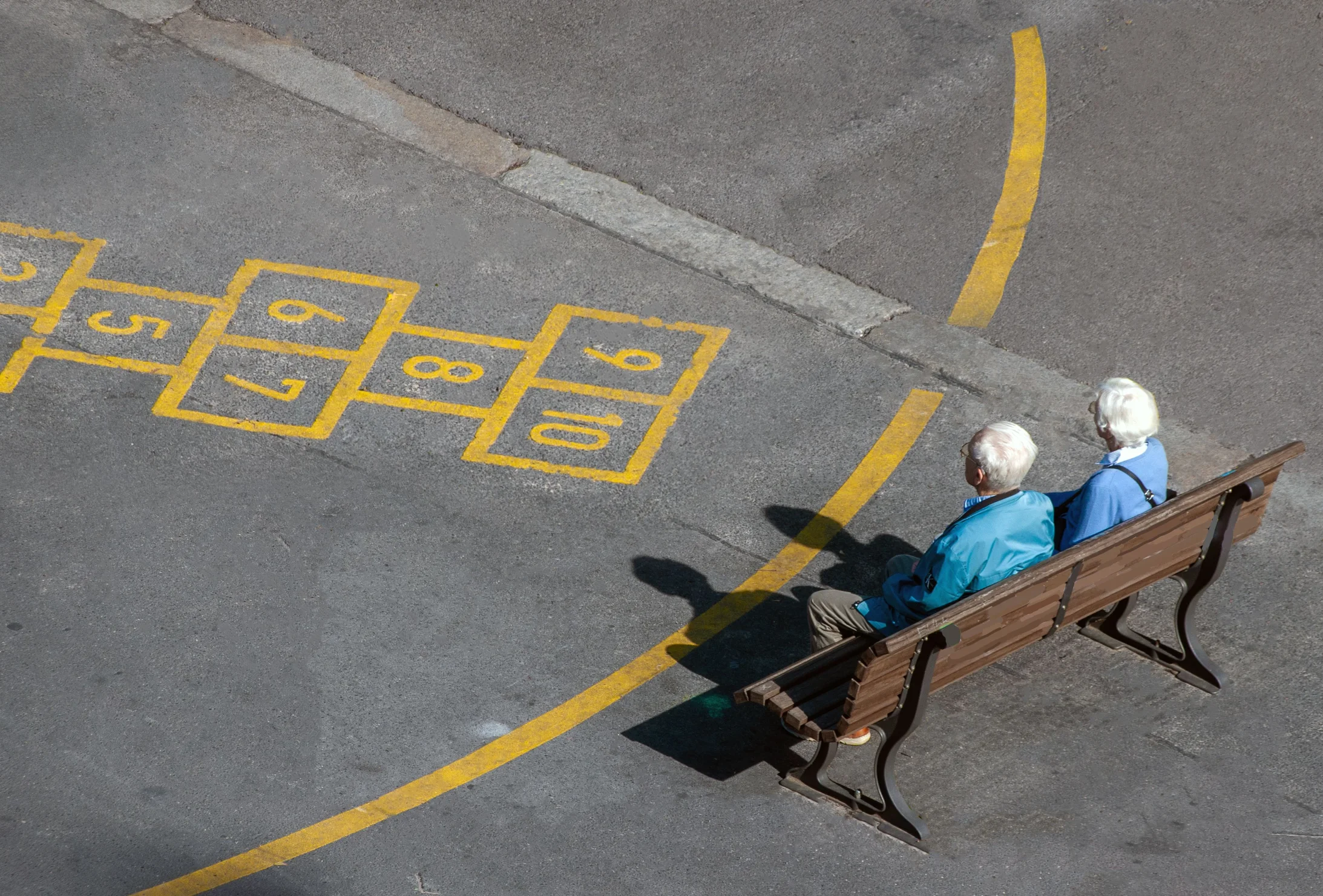 An elderly couple enjoys a sunny day on a park bench next to a hopscotch game.