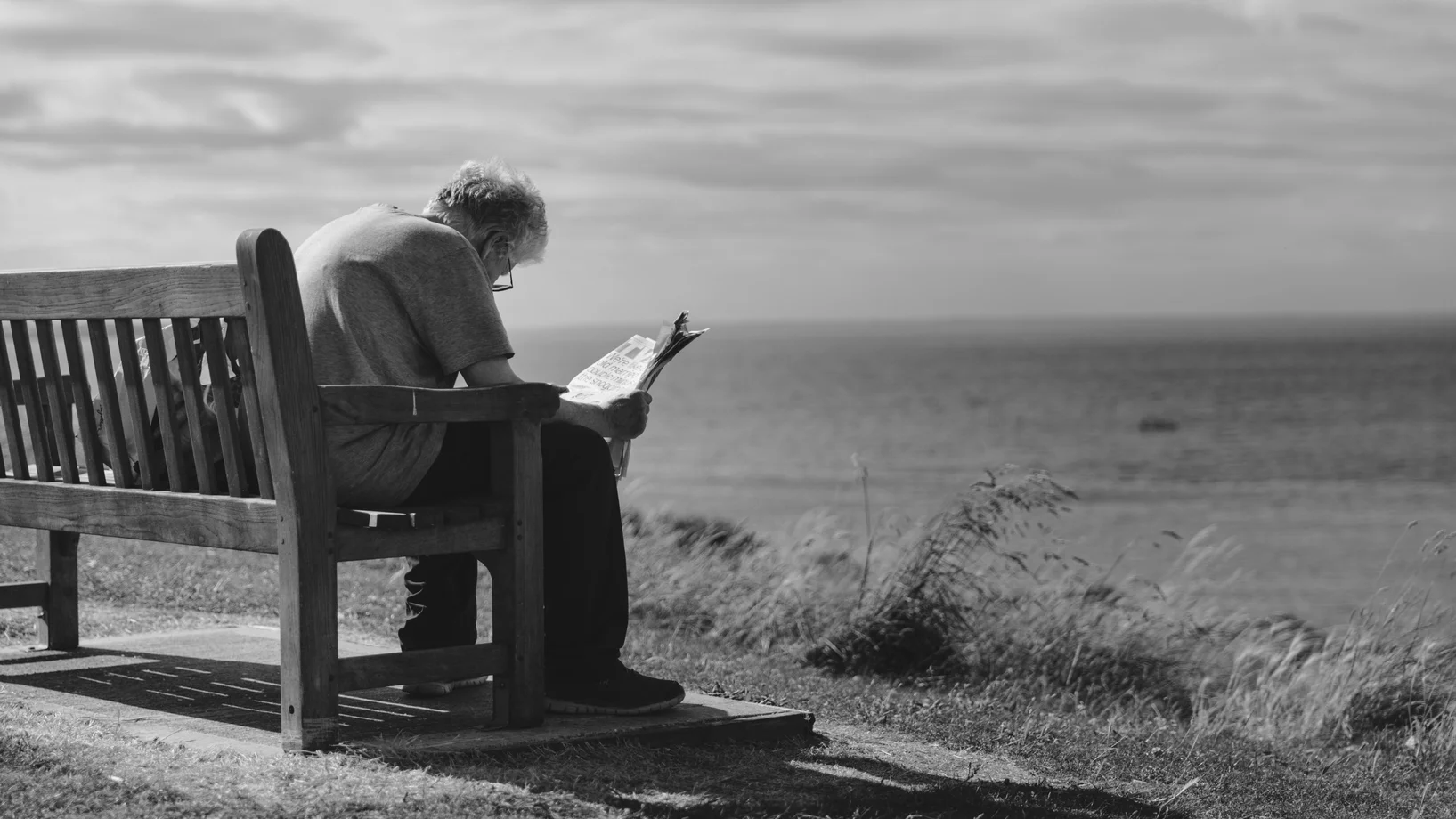 A contemplative scene of an elderly man reading a newspaper on a seaside bench with cloudy skies.