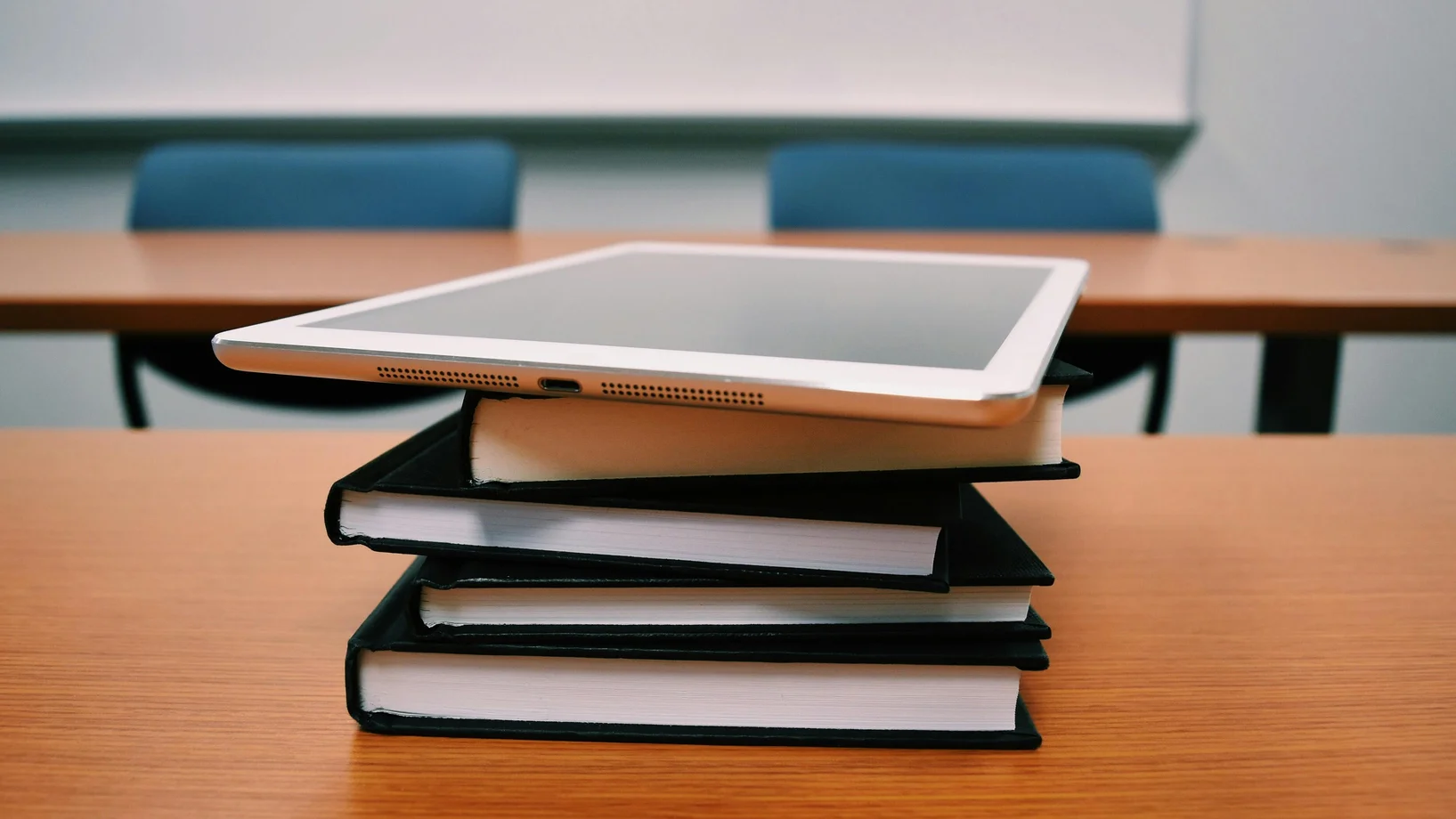 A tablet rests on top of a stack of books in an empty classroom, illustrating modern education.