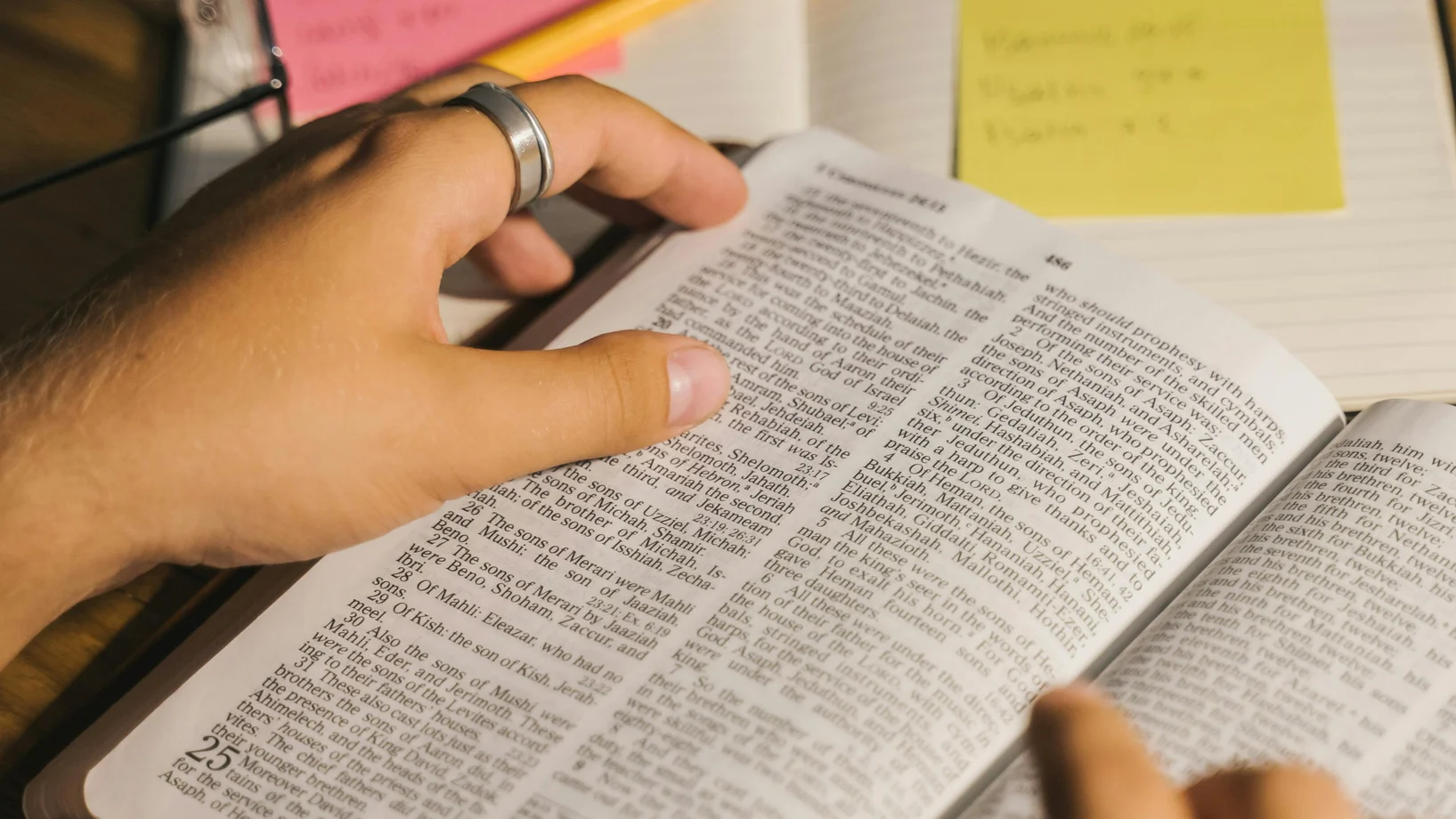 Close-up of hands holding and reading a Bible with study notes and a pencil on a desk.