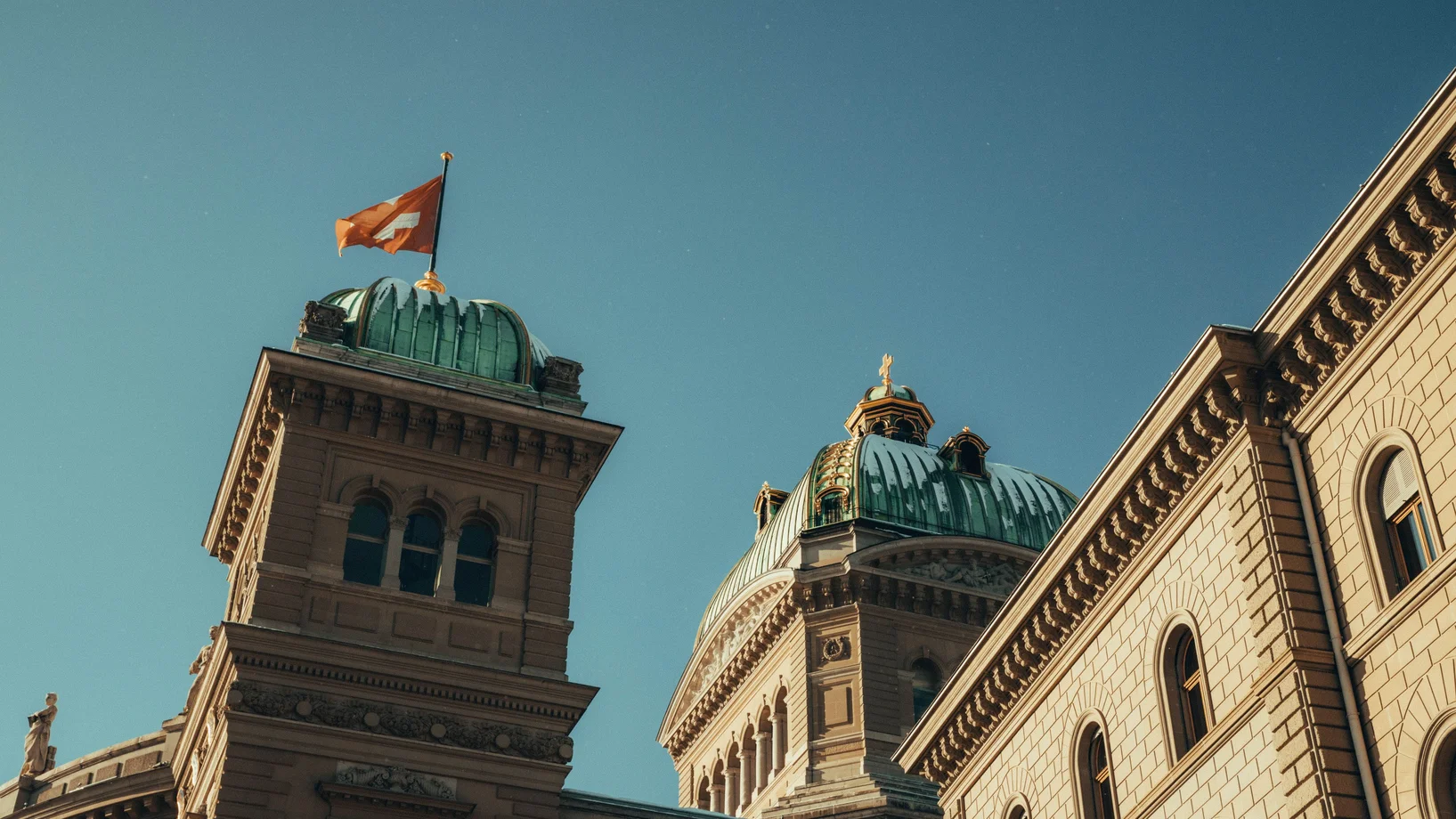 Low-angle view of the Swiss Parliament Building with flag in Berne, Switzerland under a clear blue sky.