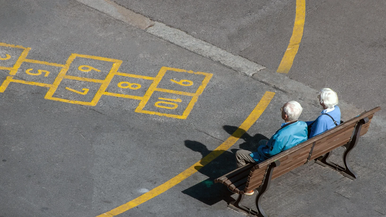 An elderly couple enjoys a sunny day on a park bench next to a hopscotch game.
